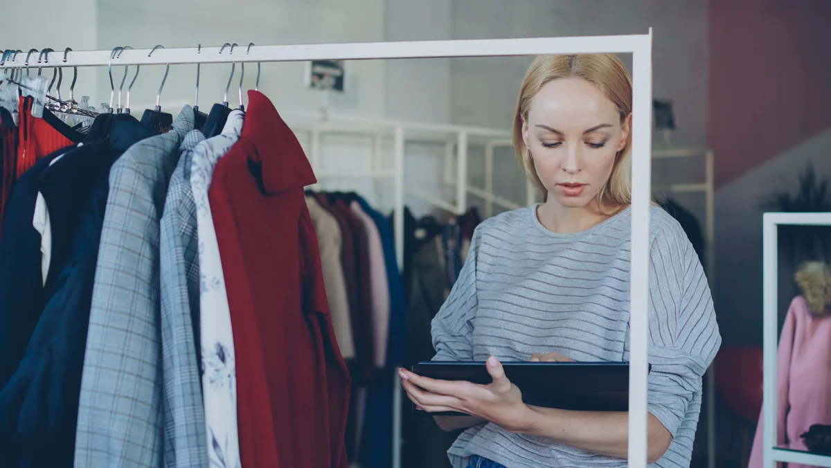 Employée de magasin consultant une tablette devant des portants de vêtements.